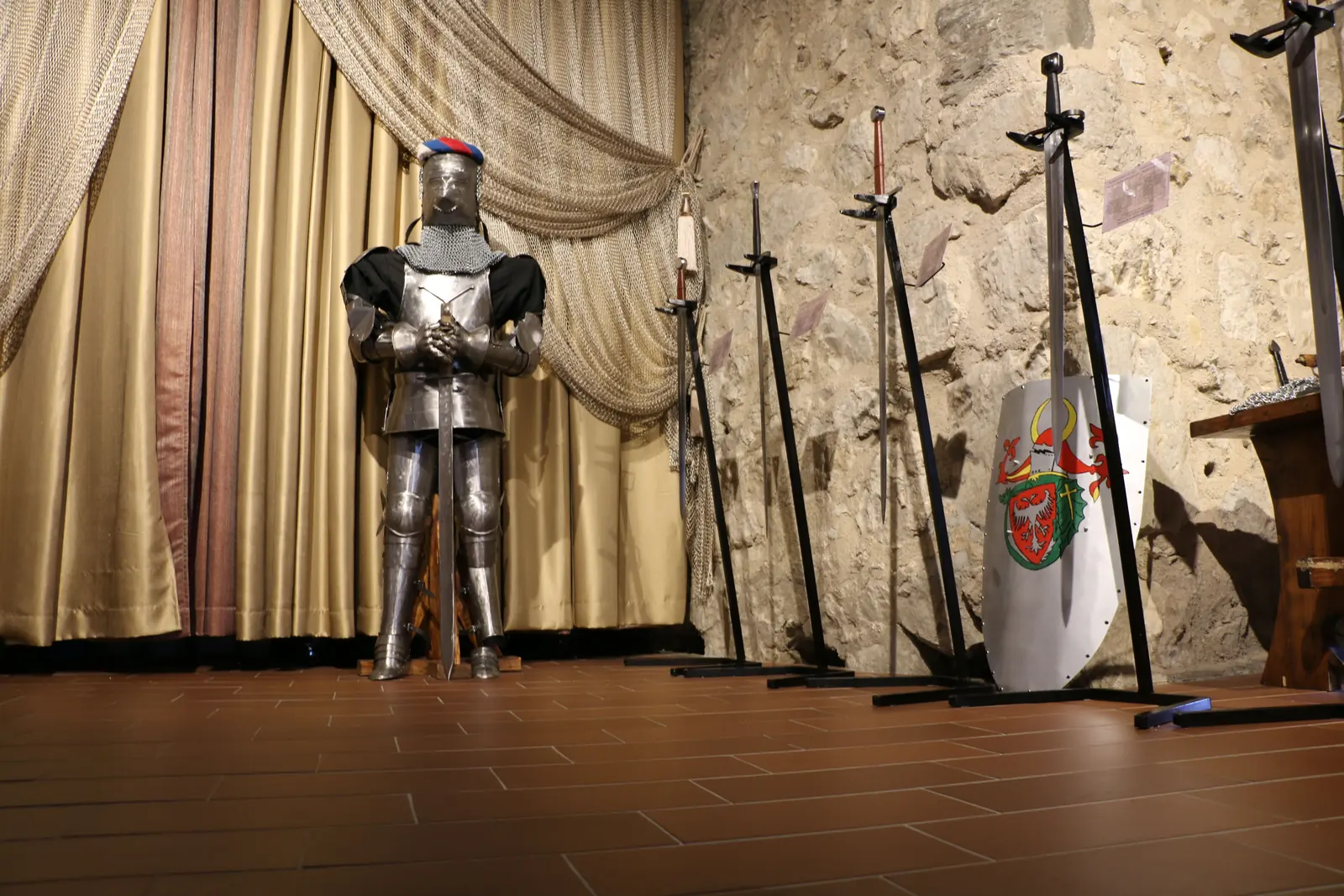 A suit of armour stands beside racked swords and a heraldic shield against a rough stone wall, with a gold curtain behind