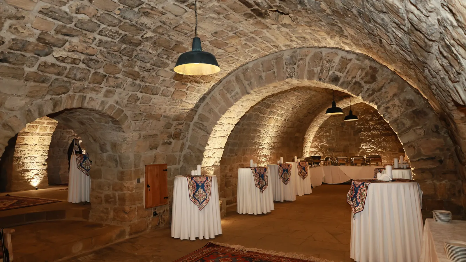 A vaulted stone cellar with arched ceilings, warm wall lights and round tables dressed in white cloths, set for a banquet