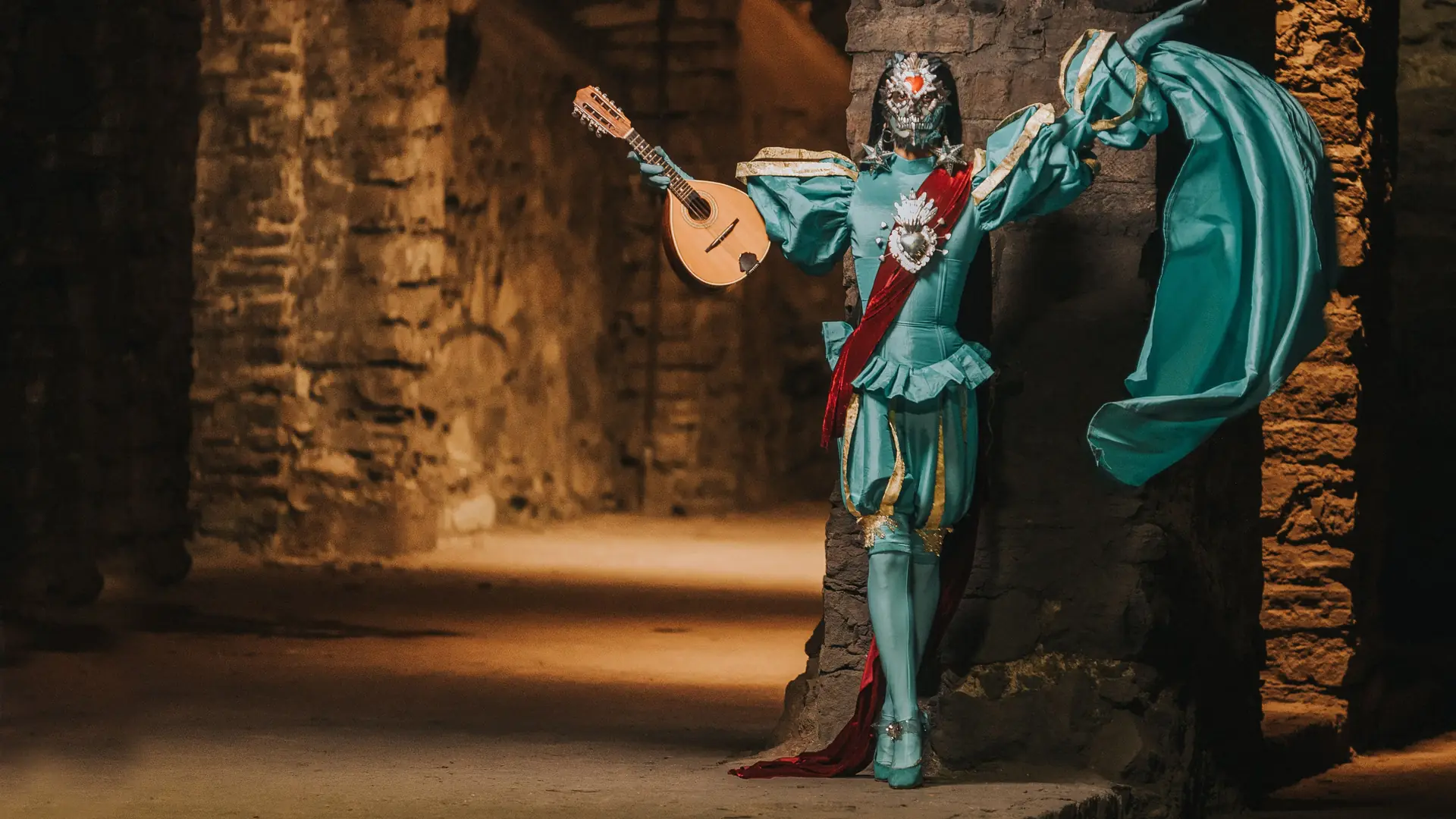 A masked minstrel in a teal doublet and red sash holds a lute and a billowing cloak against a stone pillar in a vaulted brick passage lit by warm light