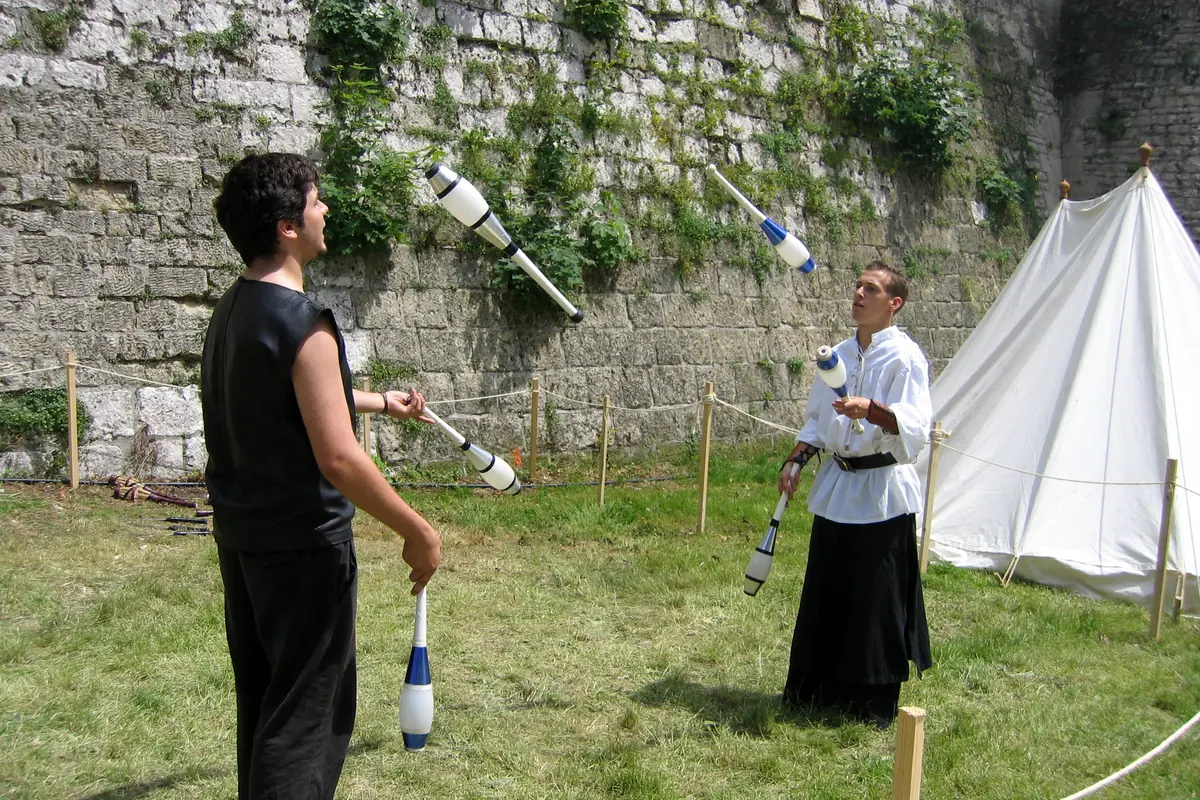 Two jugglers in medieval-style clothes throw clubs in front of a stone wall and a white tent at a fair
