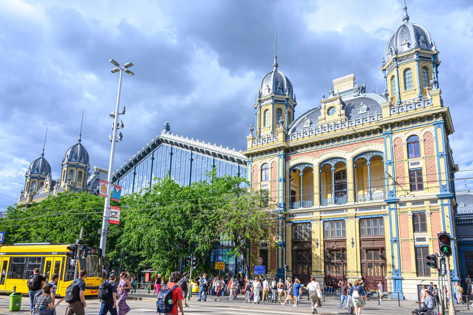 The ornate yellow and blue facade of Nyugati station in Budapest with its iron and glass hall, a yellow tram and crowds on the pavement